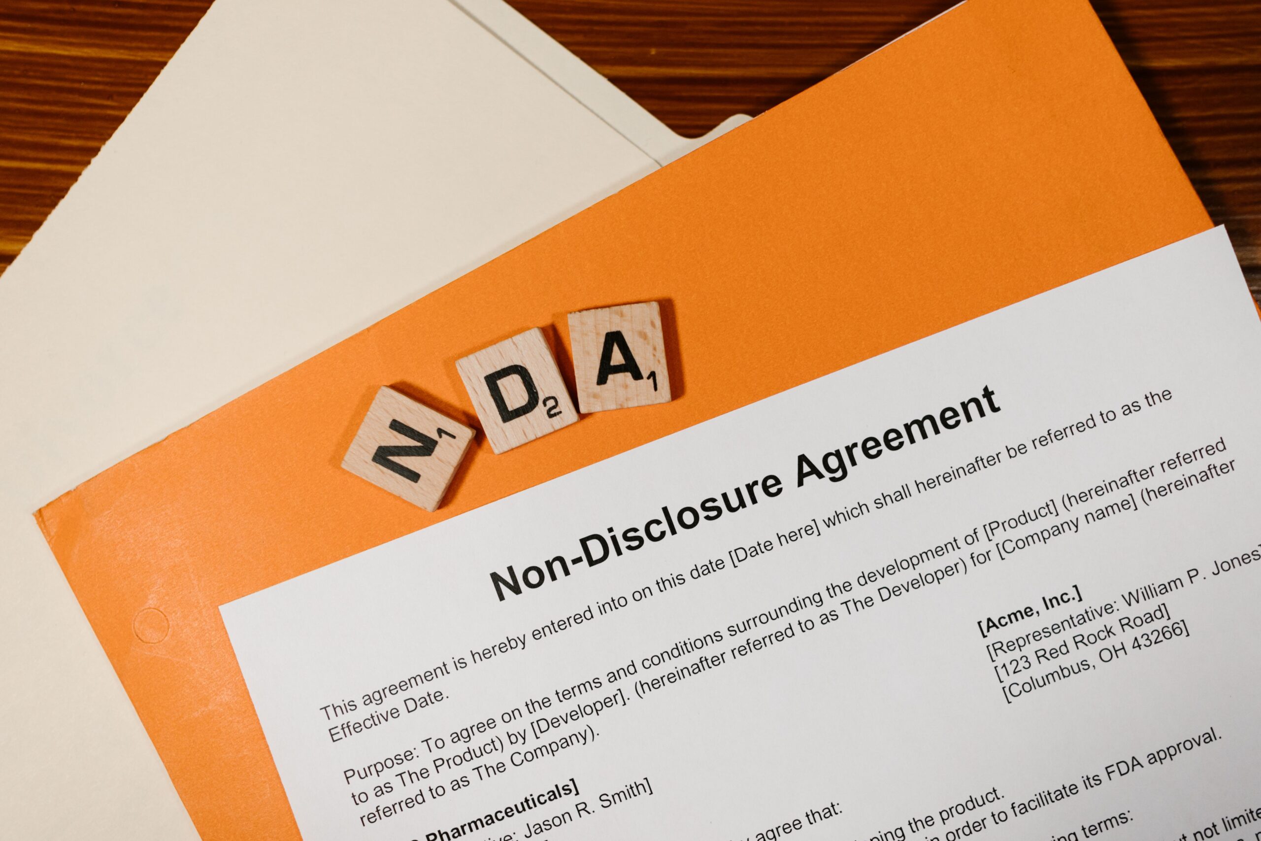 Wooden letter tiles spelling "NDA" on an orange folder beside a printed non-disclosure agreement